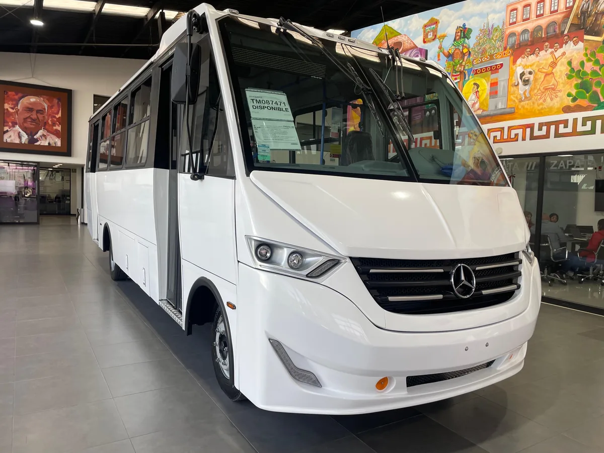 Autobús Mercedes-Benz blanco con carrocería Toreto, completo y de tres cuartos frontal, en sala de exhibición.